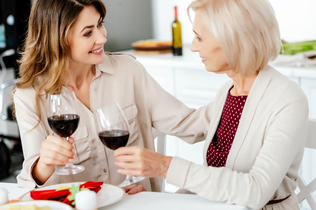 sonriente mujer adulta tintineando por copas de vino con la madre mayor en la mesa servida para la celebración de Acción de Gracias
  - Foto, Imagen