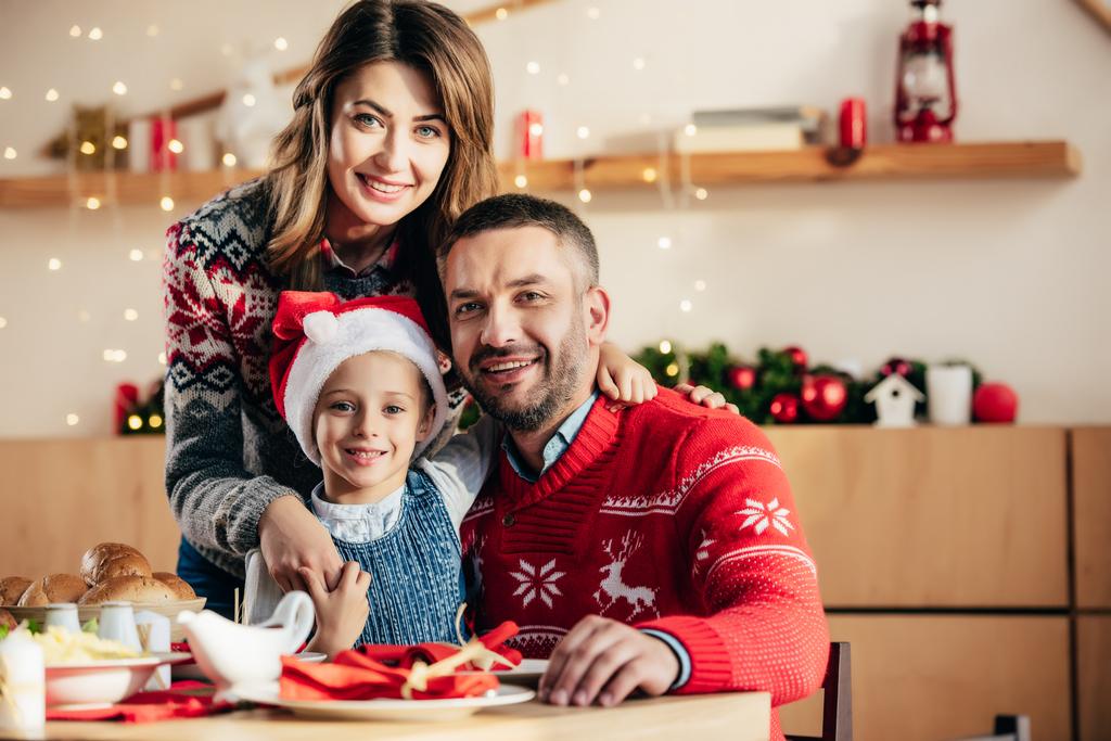 Porträt einer glücklichen Familie mit Tochter in Weihnachtsmütze am Tisch mit Festtagsessen  - Foto, Bild