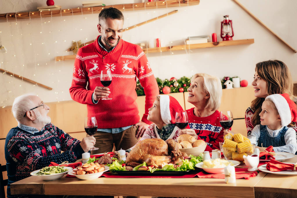 ワインのガラスを保持していると、自宅で家族とクリスマスのお祝い中にトーストを作る幸せな大人の男 - 写真・画像