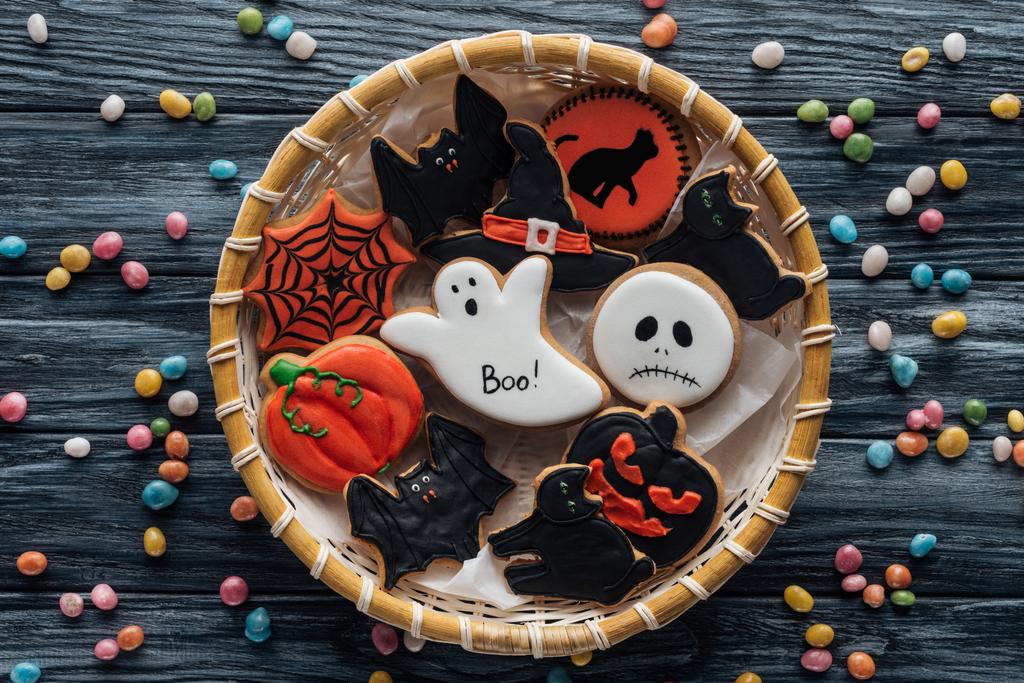 Ansicht von Weidenkorb mit hausgemachten gruseligen Halloween-Plätzchen umgeben von Bonbons auf Holztisch  - Foto, Bild