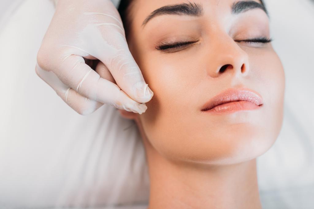 Teilansicht des Kosmetikers, der während der Akupunkturtherapie im Wellness-Salon Nadel auf das Gesicht der Frau legt - Foto, Bild