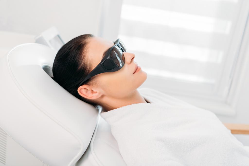 mujer en gafas protectoras consiguiendo depilación láser hecha por cosmetólogo en salón de spa
 - Foto, Imagen