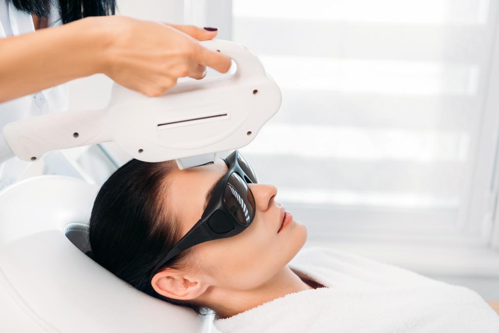 mujer en gafas protectoras consiguiendo depilación láser hecha por cosmetólogo en salón de spa
 - Foto, imagen