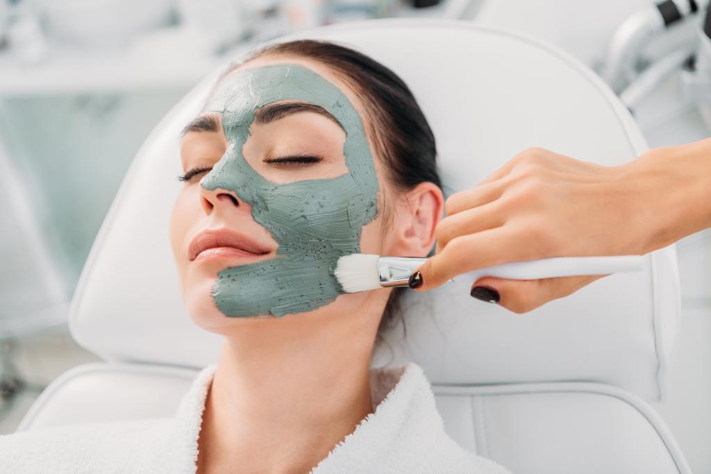частичный вид косметолога, надевающего глиняную маску на женское лицо в спа салоне
 - Фото, изображение