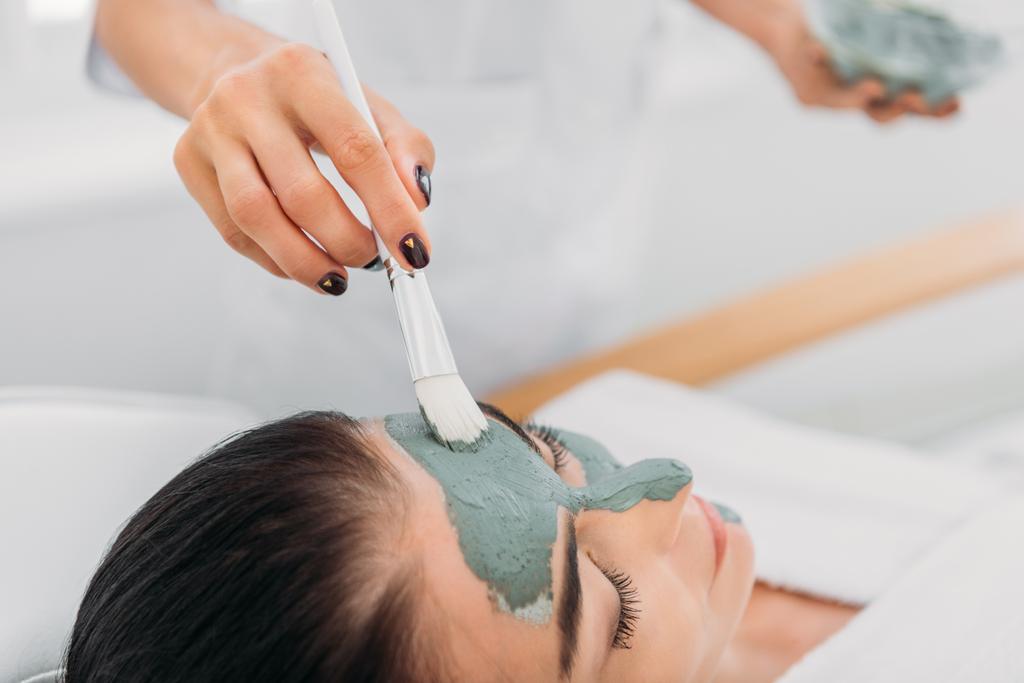 Teilansicht des Kosmetologen, der im Wellness-Salon Tonmaske mit Pinsel auf das weibliche Gesicht aufträgt - Foto, Bild