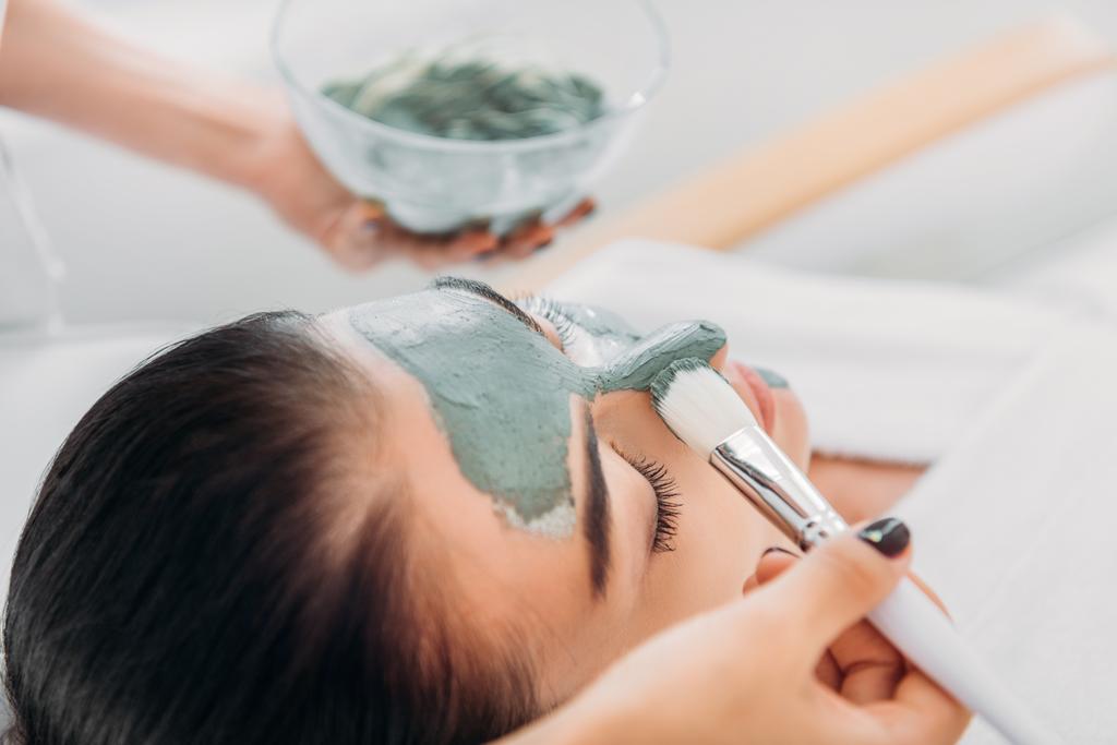 częściowy widok kosmetolog stosowania maski gliny z pędzla na twarz kobiety w salonie spa - Zdjęcie, obraz