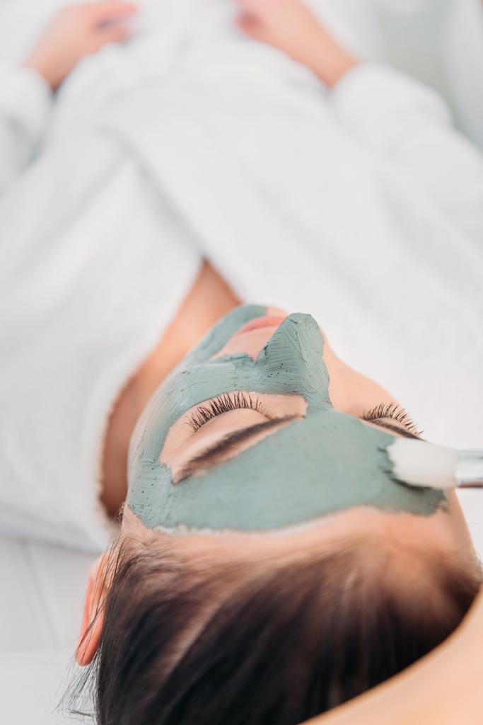 częściowy widok kosmetolog stosowania maski gliny na twarz kobiety w salonie spa - Zdjęcie, obraz