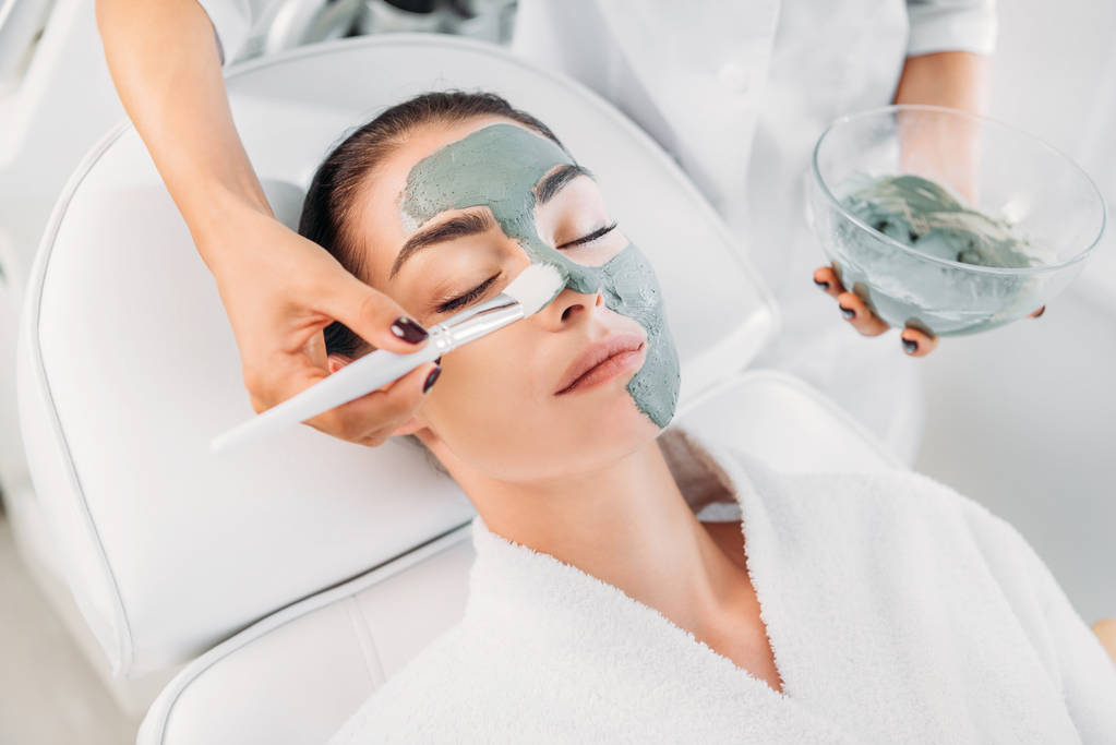 gedeeltelijke weergave van schoonheidsspecialist klei masker met borstel toe te passen op vrouwelijke gezicht in spa salon - Foto, afbeelding