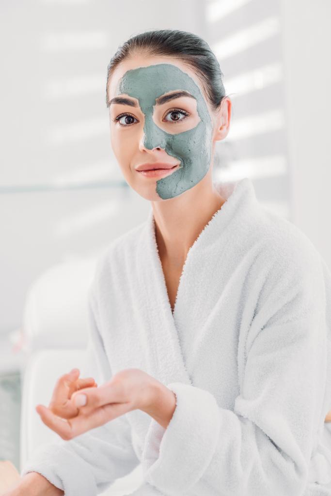 portrait of beautiful woman with clay mask on face in white bathrobe in spa salon  - Photo, Image