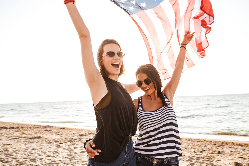 Εικόνα των ευτυχισμένων γυναικών φίλους σε εξωτερικούς χώρους στην παραλία κρατώντας τη σημαία των ΗΠΑ διασκεδάζοντας. - Φωτογραφία, εικόνα