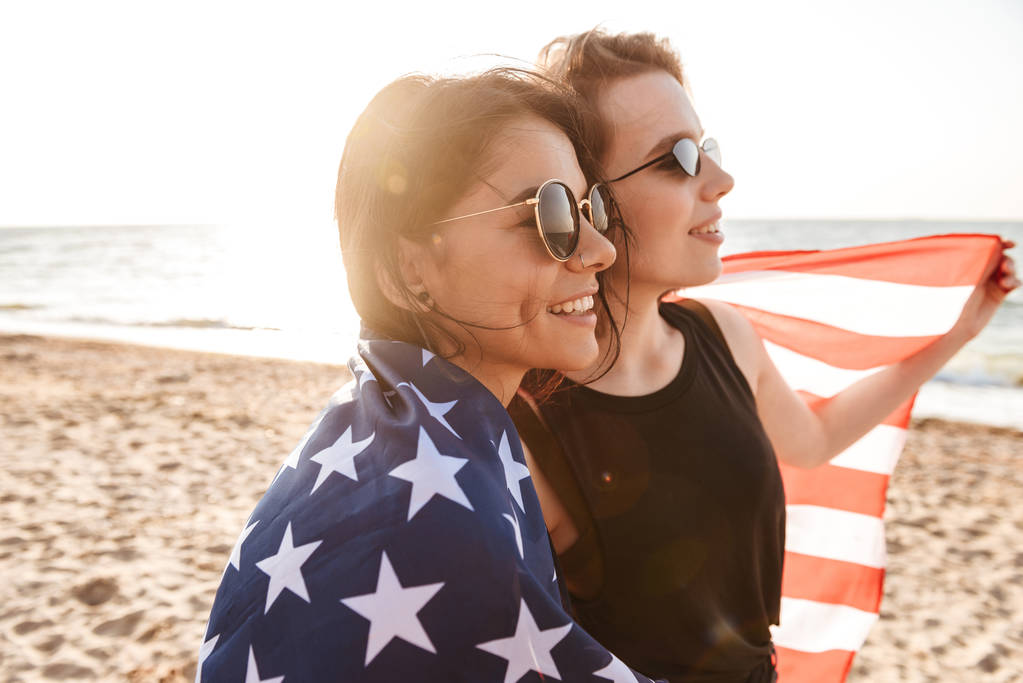 Image de jolies amies excitées à l'extérieur sur la plage tenant le drapeau des États-Unis s'amusant
. - Photo, image