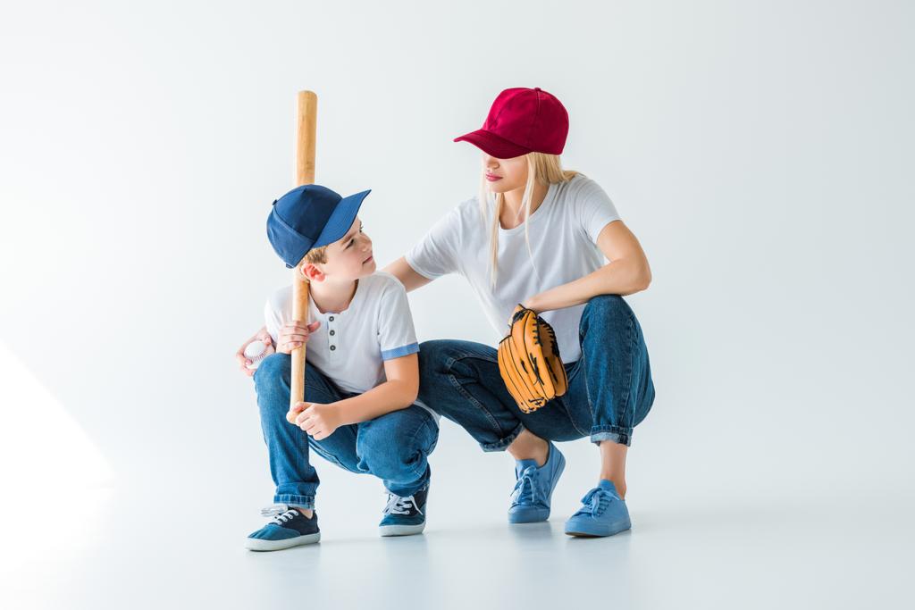 Máma a syn v podřepu s baseballovou pálkou a rukavice na bílém pozadí a při pohledu na sebe - Fotografie, Obrázek