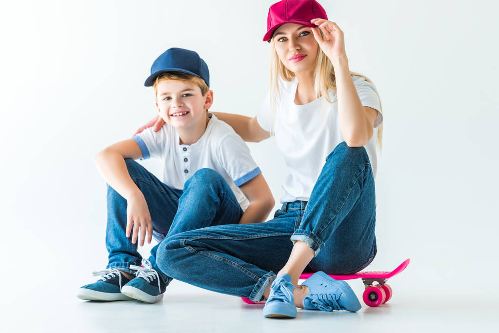 mother and son in caps sitting on skates, hugging and looking at camera on white - Photo, Image