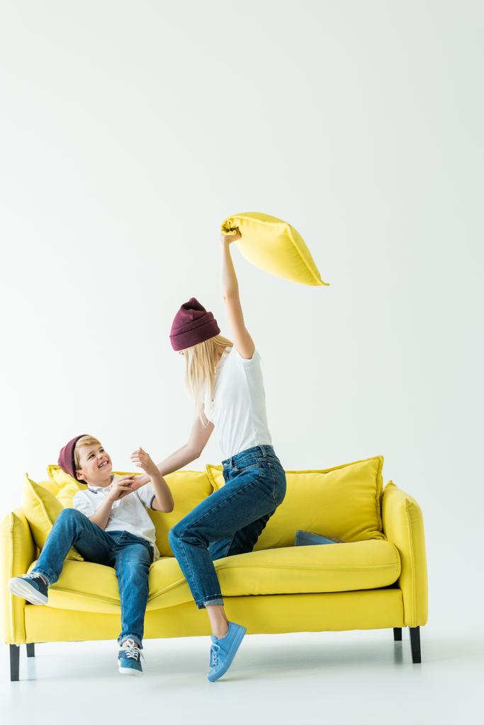 μητέρα διασκεδάζοντας και ξυλοδαρμό γιο με μαξιλάρι σε κίτρινο καναπέ σε λευκό - Φωτογραφία, εικόνα