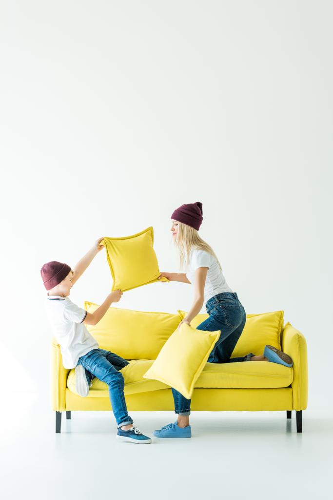 Mutter und Sohn in weinroten Hüten haben Spaß und kämpfen mit Kissen auf gelbem Sofa auf weißem - Foto, Bild
