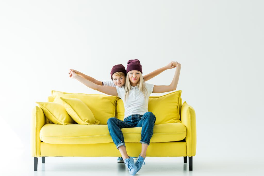 mother and son in burgundy hats pretending flying on yellow sofa on white - Photo, Image