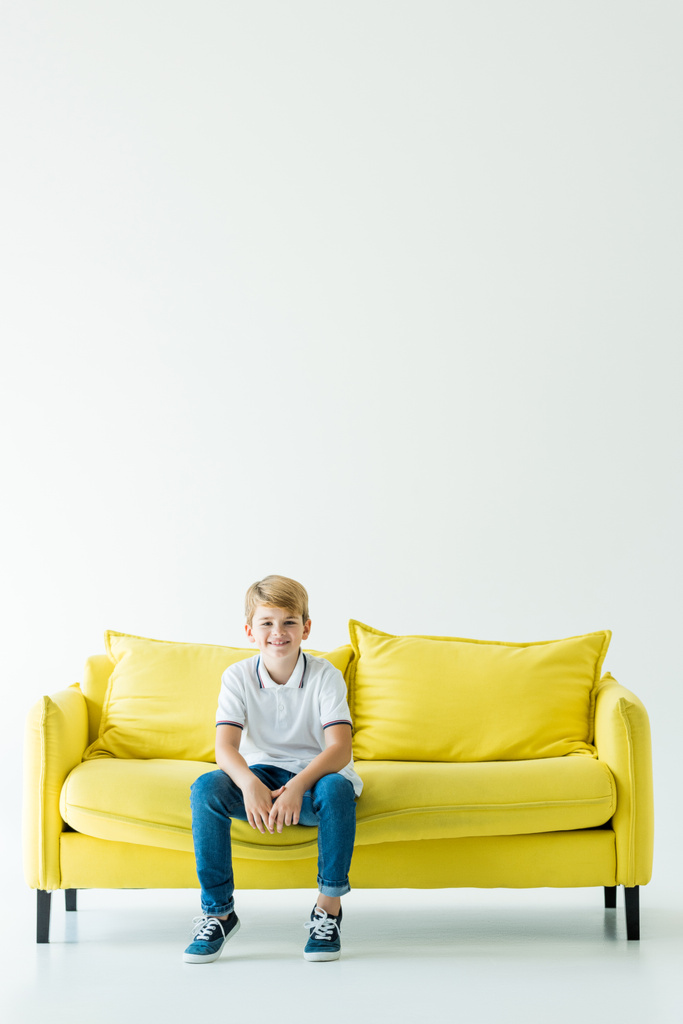 χαμογελώντας αξιολάτρευτο παιδί κάθεται στο κίτρινο καναπέ και να βλέπουν τα φωτογραφικών μηχανών σε λευκό - Φωτογραφία, εικόνα