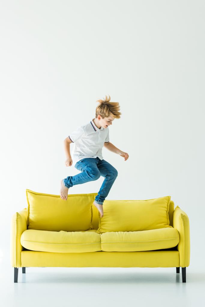 αξιολάτρευτο παιδί σε casual ρούχα άλμα σε κίτρινο καναπέ σε λευκό - Φωτογραφία, εικόνα