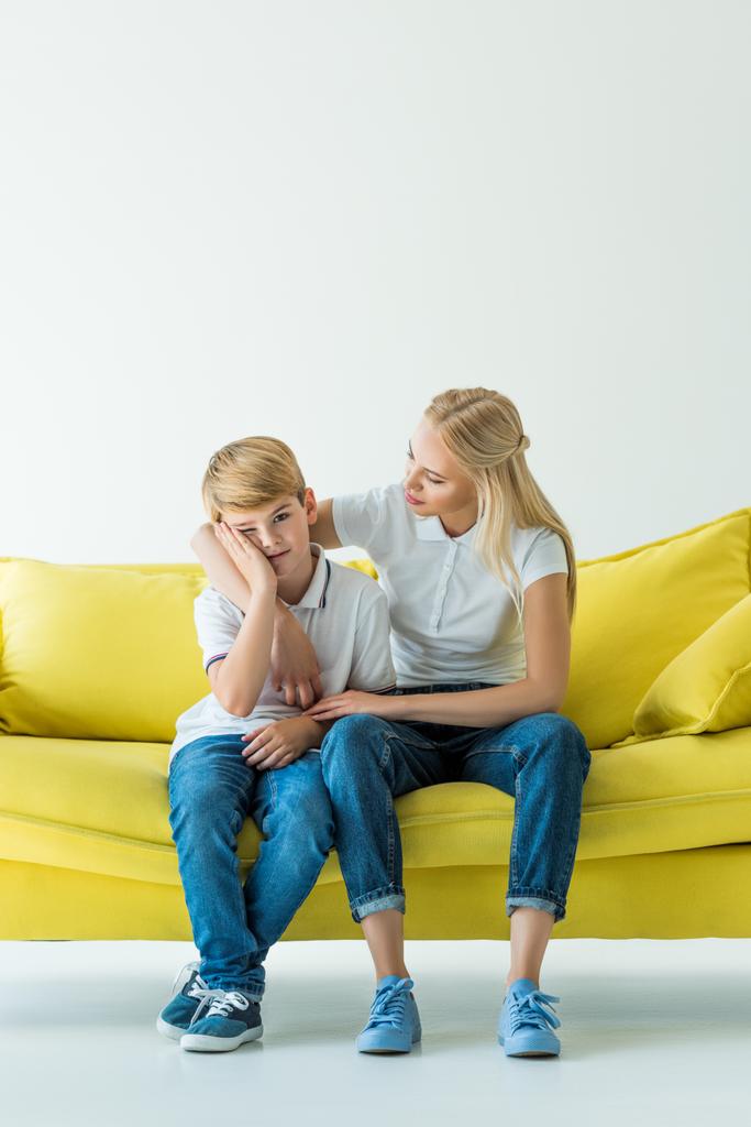 Mutter umarmt verärgerten Sohn auf gelbem Sofa auf weißem Grund - Foto, Bild
