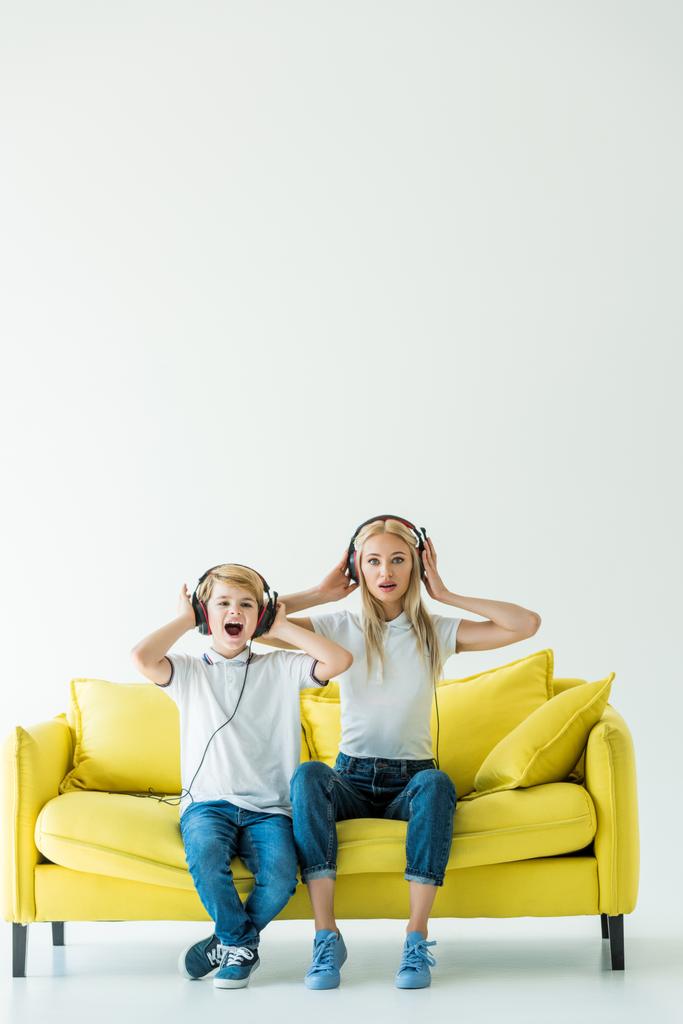geschokt moeder en zoon luisteren muziek met koptelefoon op gele sofa op wit - Foto, afbeelding