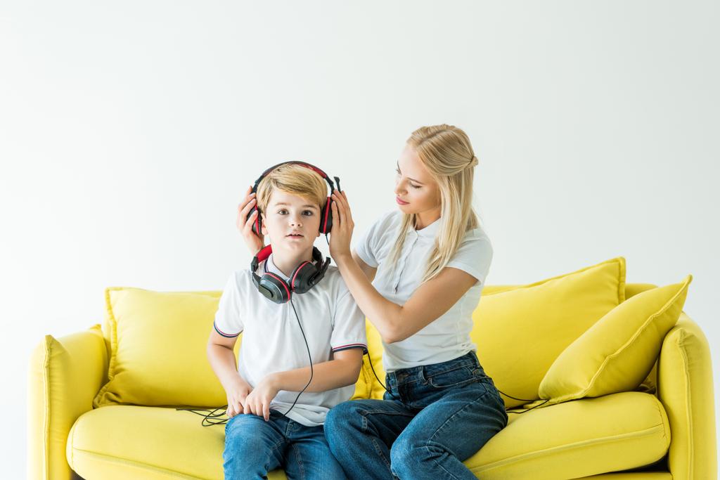 Mutter trägt Kopfhörer auf Sohn auf gelbem Sofa isoliert auf weißem - Foto, Bild