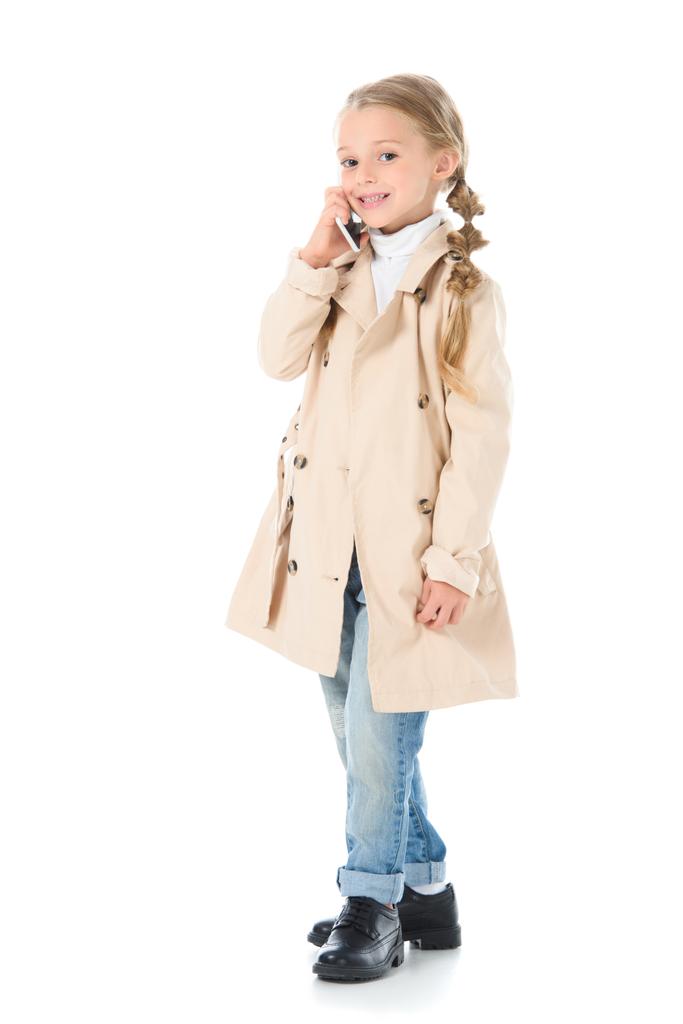 Gelukkig schattig kind in beige jas praten op smartphone, geïsoleerd op wit - Foto, afbeelding