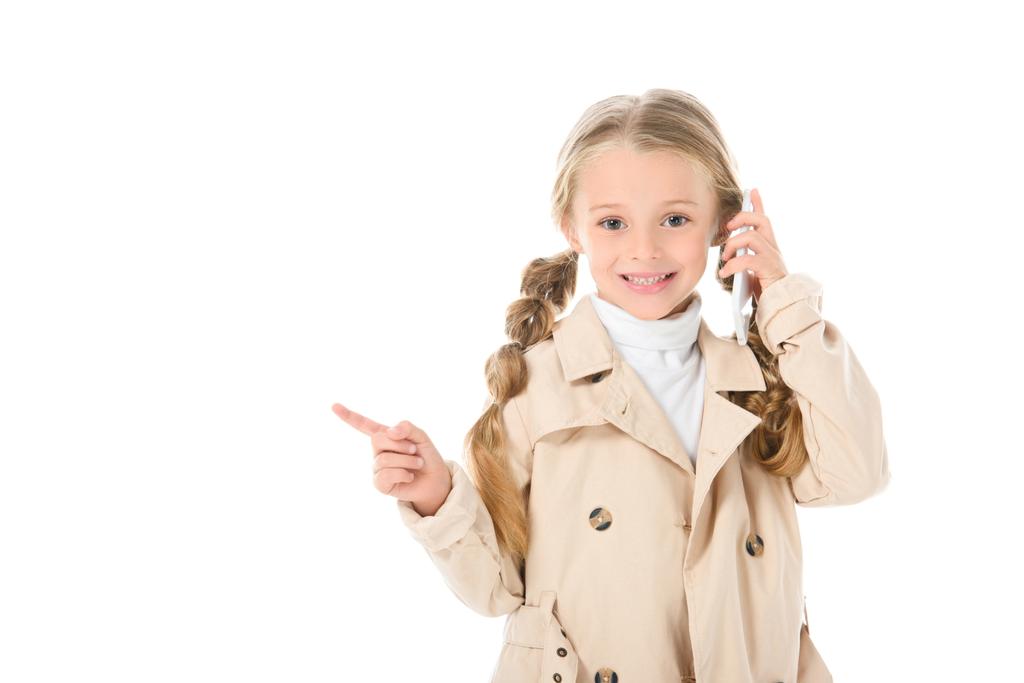 χαμογελώντας παιδιού σε μπεζ παλτό μιλώντας στο smartphone και δείχνει κάτι, απομονώνονται σε λευκό - Φωτογραφία, εικόνα