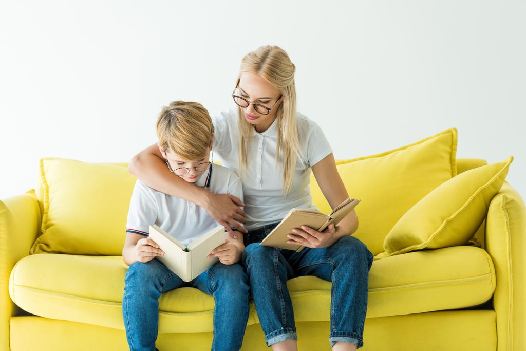 мать в очках обнимает сына, пока он читает книгу на желтом диване, изолированном на белом
 - Фото, изображение