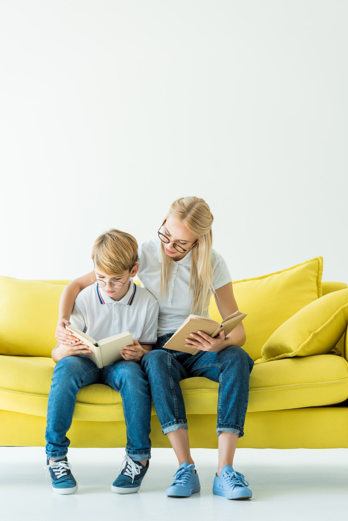 мать обнимает сына, пока он читает книгу на жёлтом диване на белом
 - Фото, изображение