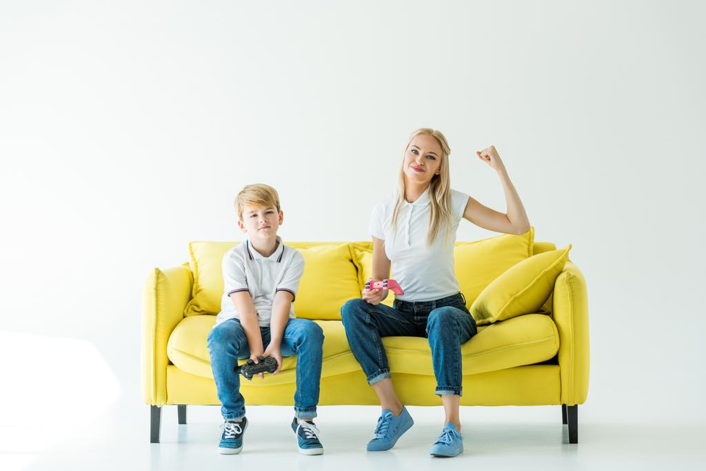 Glückliche Mutter zeigt Ja-Geste nach gewonnenem Videospiel auf gelbem Sofa auf weißem Grund - Foto, Bild