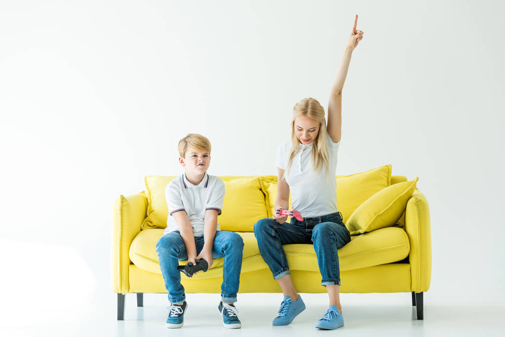 μητέρα δείχνει ένα χέρι επάνω μετά από το νικηφόρο παιχνίδι βίντεο, γιος κάθεται αναστατωμένος στο κίτρινο καναπέ σε λευκό - Φωτογραφία, εικόνα