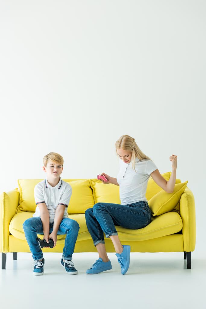 mother showing yes gesture after winning video game, son sitting upset on yellow sofa on white - Photo, Image