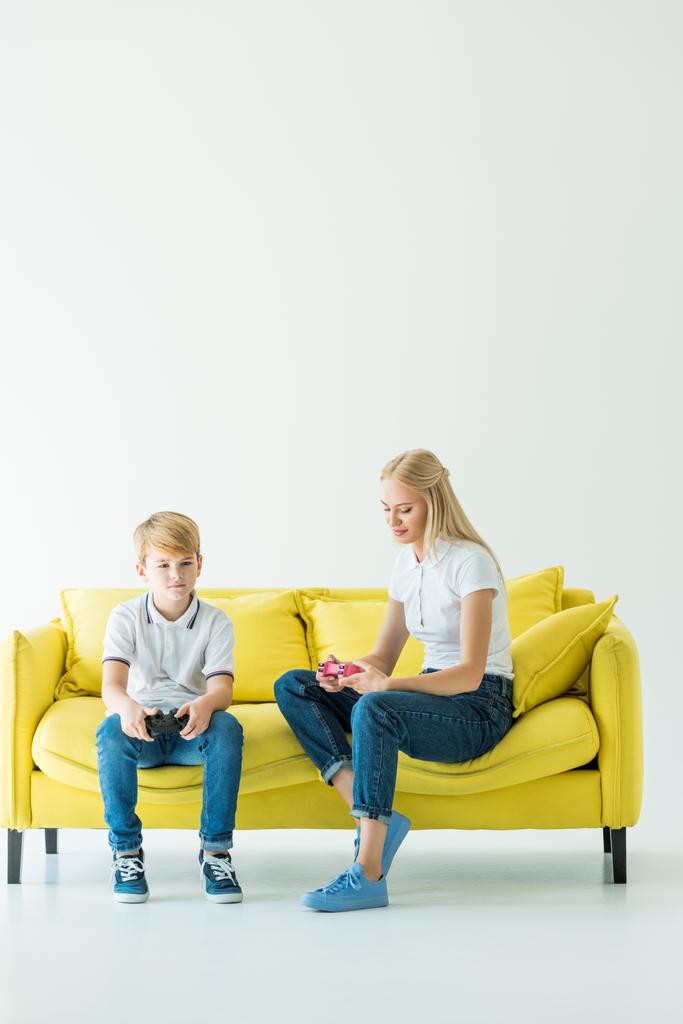 mother and son sitting with gamepads on yellow sofa on white - Photo, Image