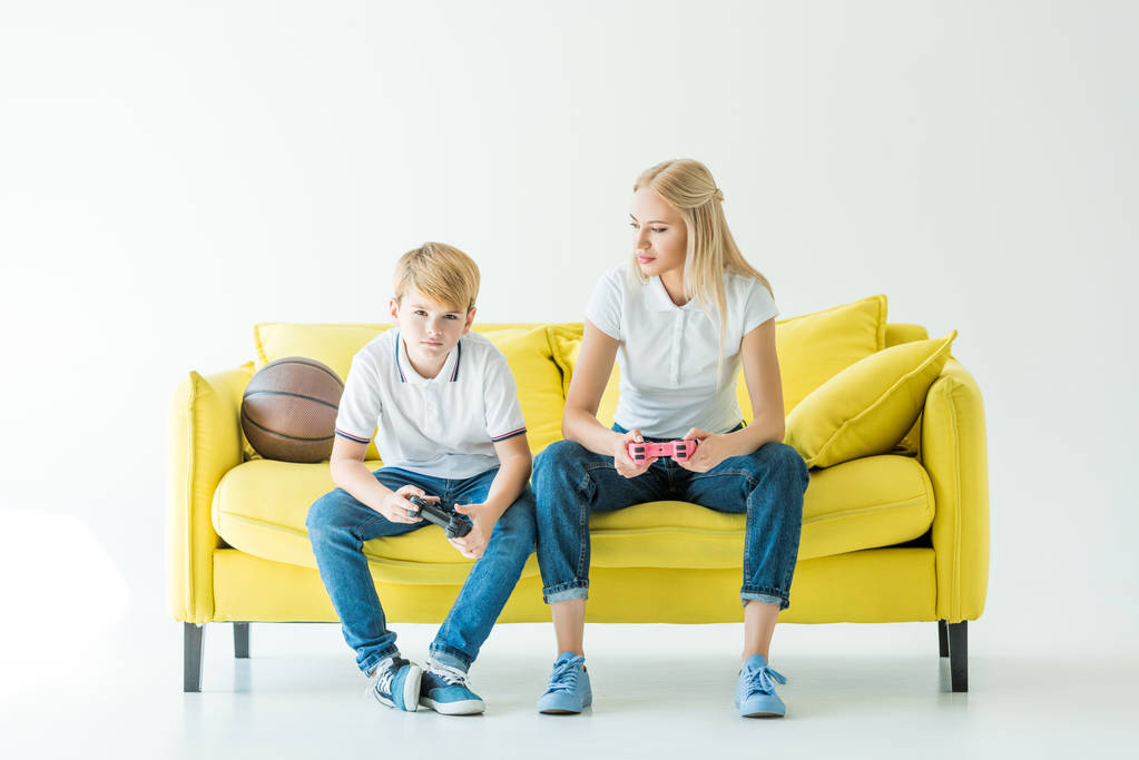 moeder zoon kijken tijdens het afspelen van video game met gamepads, basketbal bal op gele Bank - Foto, afbeelding