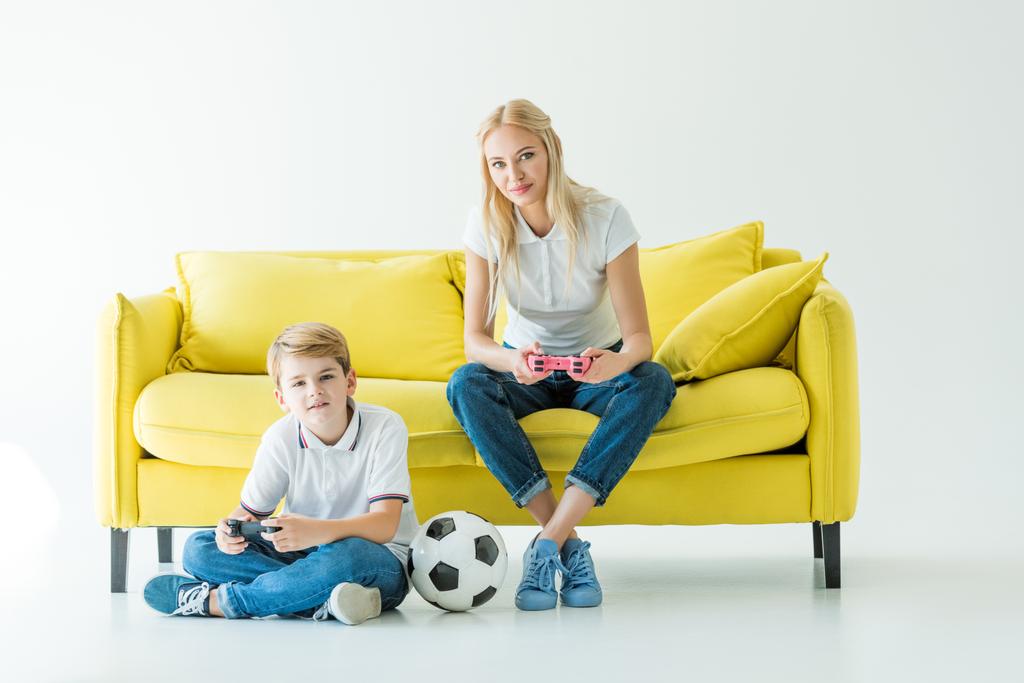 mother and son playing video game on yellow sofa on white, football ball on floor - Photo, Image
