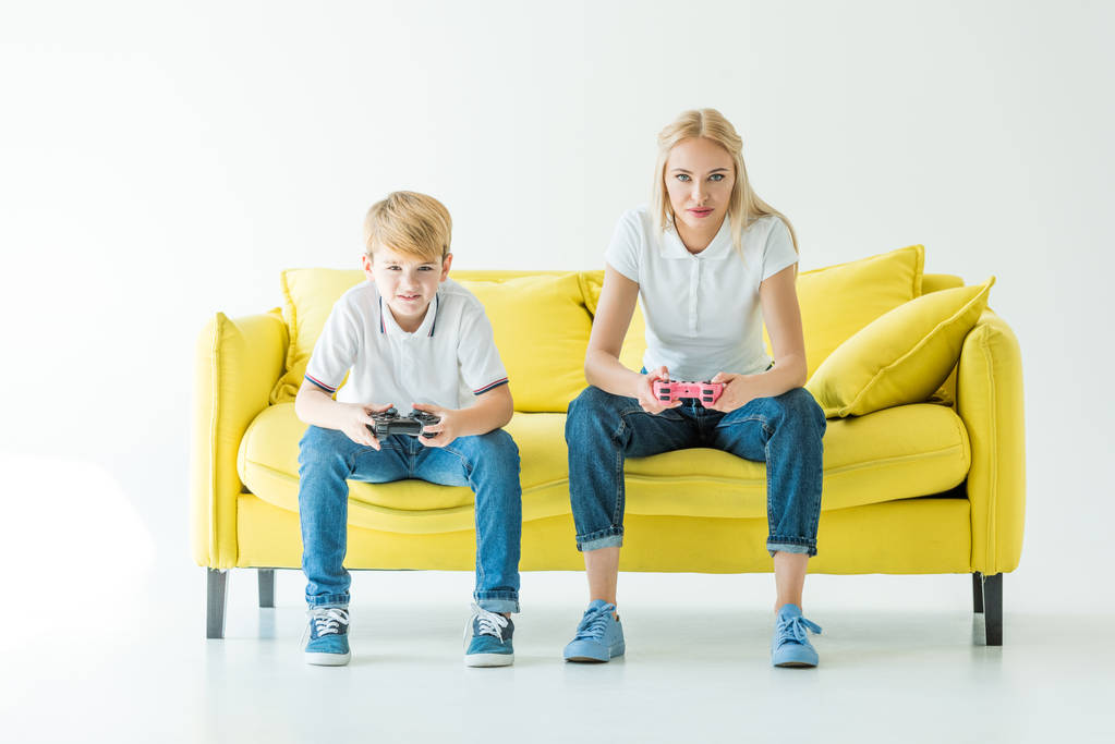 serious mother and son playing video game on yellow sofa on white - Photo, Image