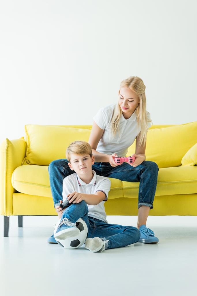 Äiti katselee poikaa pelatessaan videopeliä keltaisella sohvalla valkoisella, jalkapallo lattialla
 - Valokuva, kuva