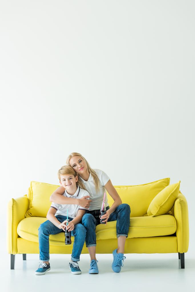 η μητέρα αγκαλιάζει γιος στον κίτρινο καναπέ και αυτοί κατέχουν σόδα σε γυάλινα μπουκάλια - Φωτογραφία, εικόνα
