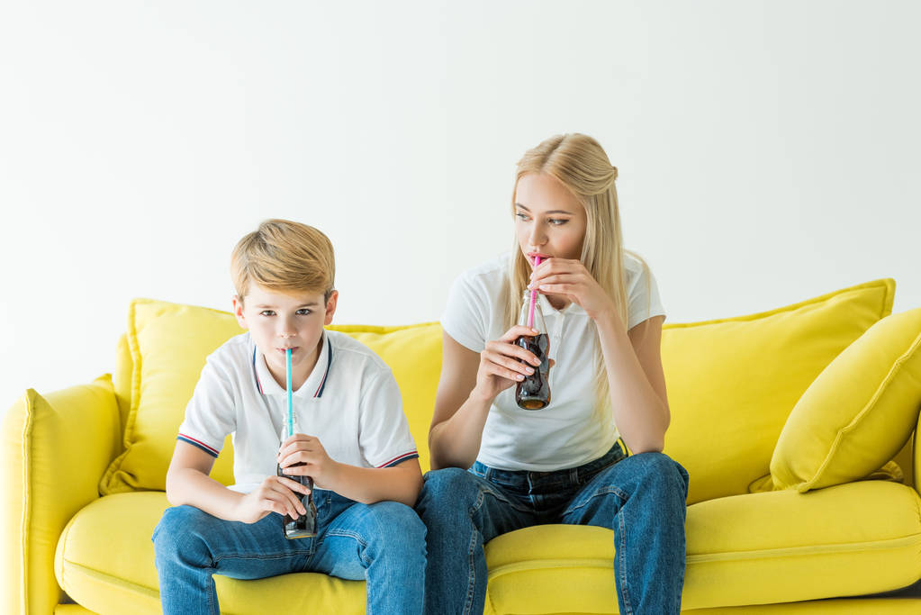 母と息子は、白で隔離黄色のソファーにストローでソーダを飲む - 写真・画像