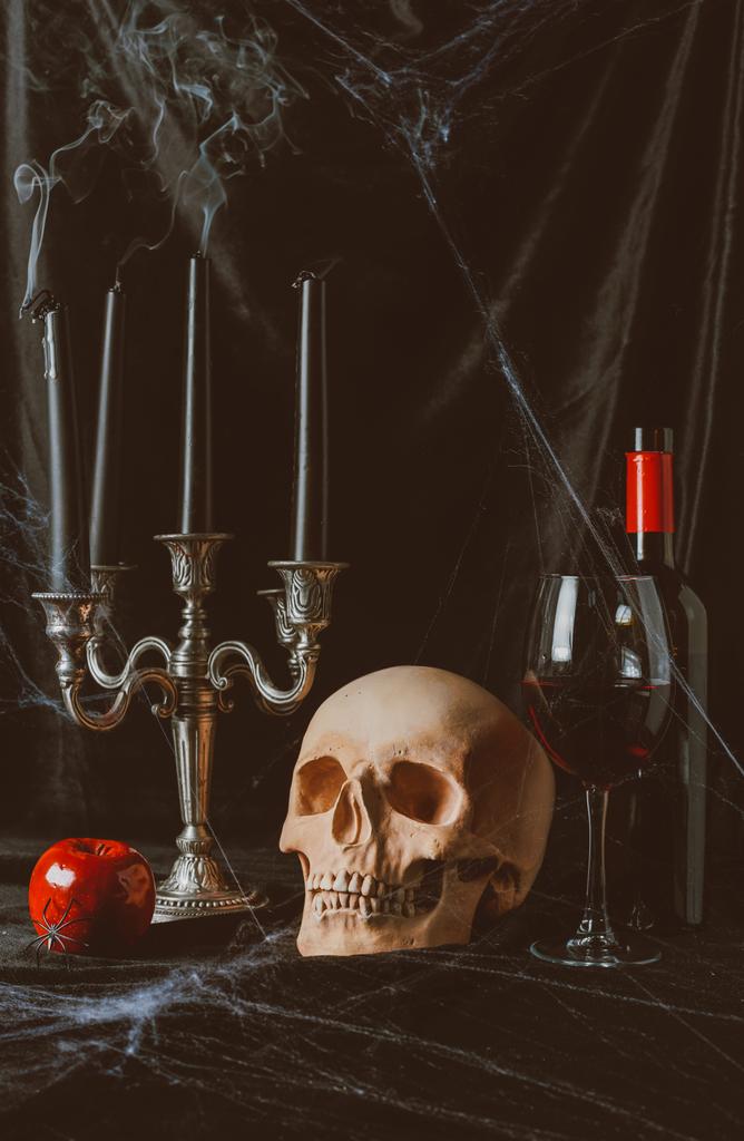 schedel, apple, rode wijn en kandelaar met rokerige kaarsen op zwarte doek met spinnenweb - Foto, afbeelding
