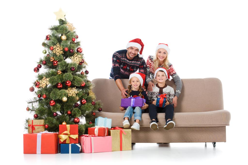 白で隔離でギフト用の箱のクリスマス ツリーの近くのソファに座っているサンタ帽子の子供と家族を笑顔で - 写真・画像