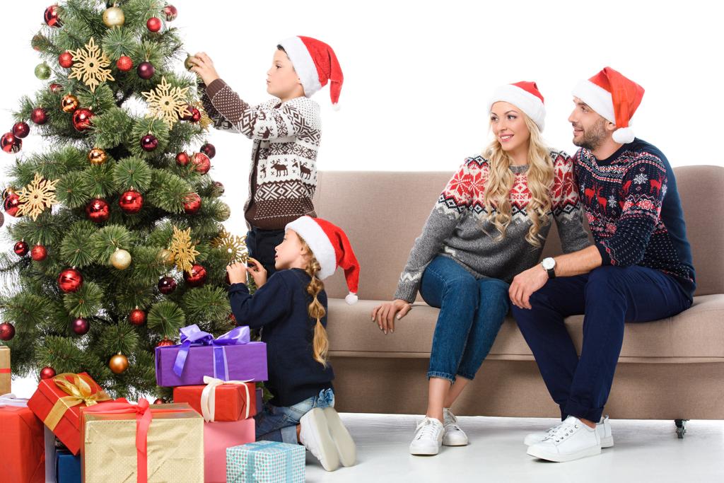Eltern mit Weihnachtsmützen betrachten Kinder, die den Weihnachtsbaum schmücken, isoliert auf weiß - Foto, Bild