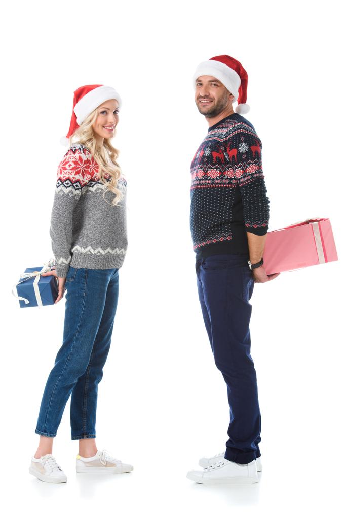 couple in christmas sweaters and santa hats hiding gift boxes, isolated on white - Photo, Image