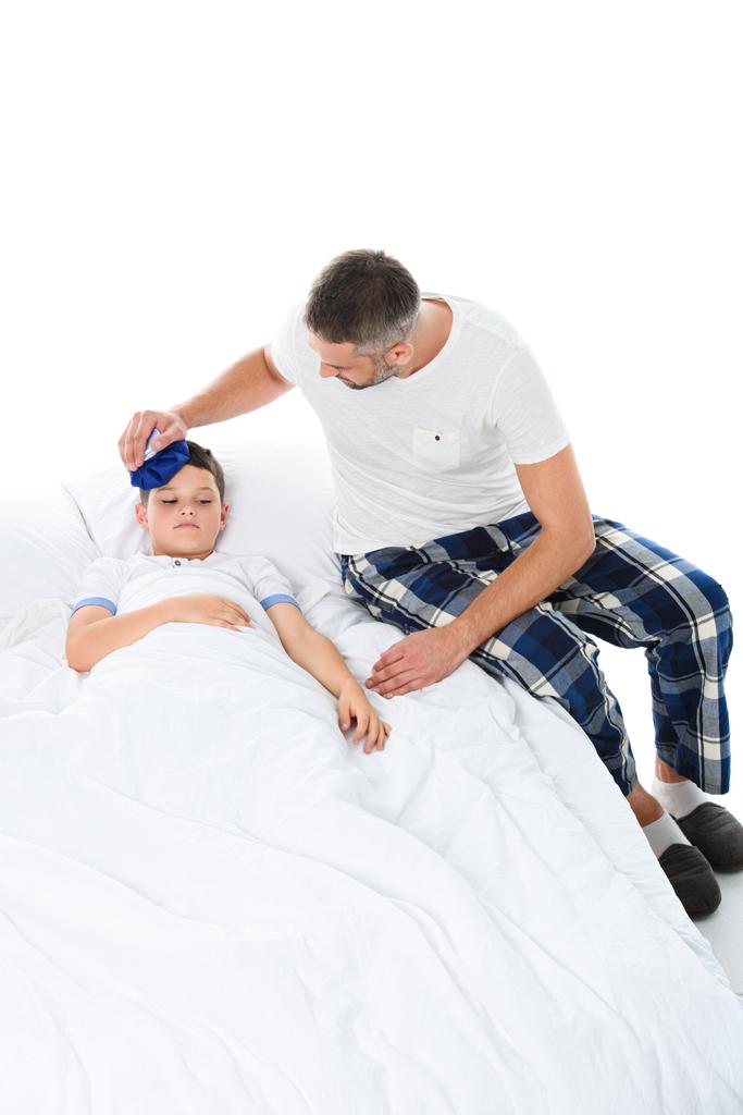 Vader pak ijs op hoofd van zieke zoon houden met temperatuur liggend in bed, geïsoleerd op wit - Foto, afbeelding
