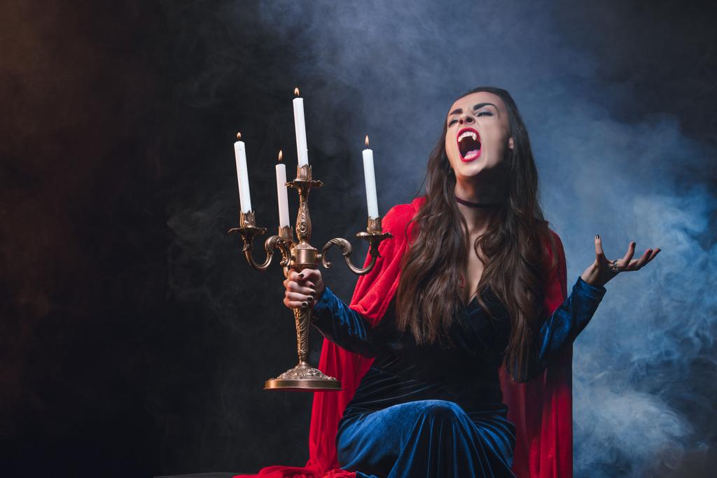 woman in red cloak holding vintage candelabrum and showing vampire fangs on dark background with smoke  - Photo, Image