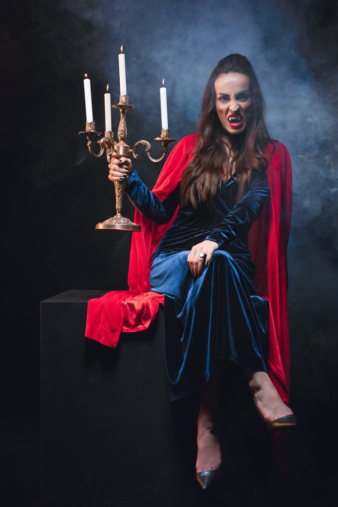 bela mulher de manto vermelho segurando candelabro retro e mostrando dentes de vampiro no fundo escuro com fumaça
  - Foto, Imagem