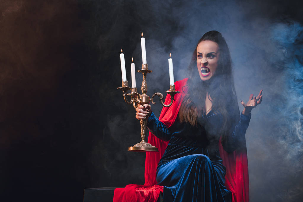 mysterie vrouw in vampier kostuum holding antieke kandelaar op donkere achtergrond met rook - Foto, afbeelding