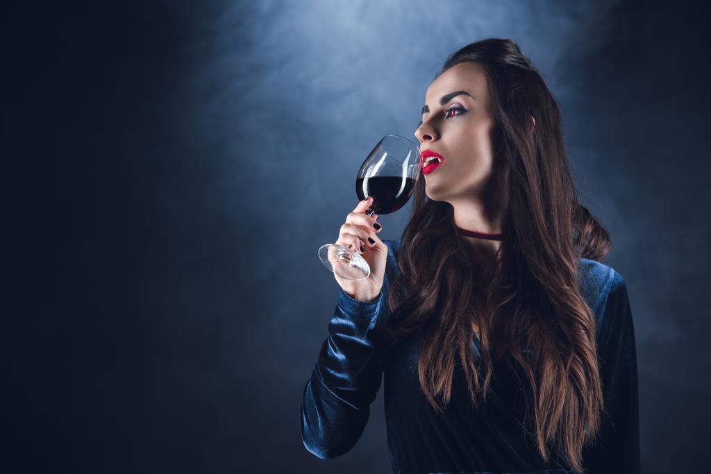 beautiful vampire drinking blood from wineglass on dark background with smoke  - Photo, Image