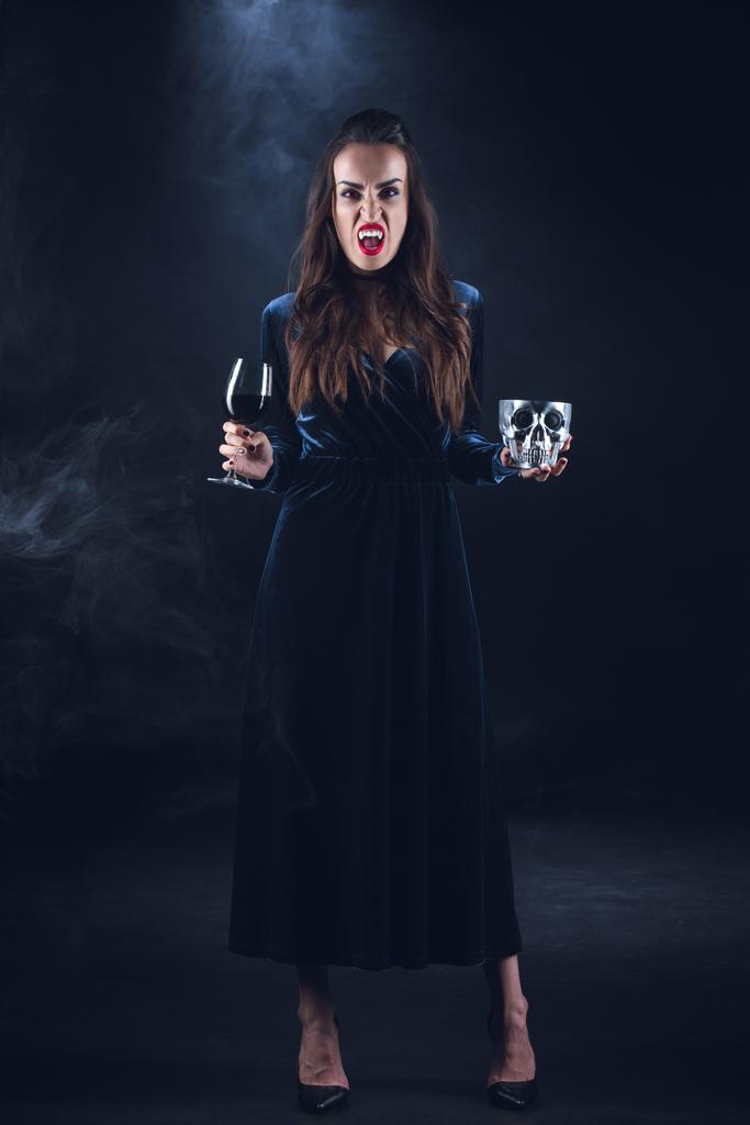 煙で暗い背景に頭蓋骨と血でワイングラスを保持している吸血鬼の女性  - 写真・画像