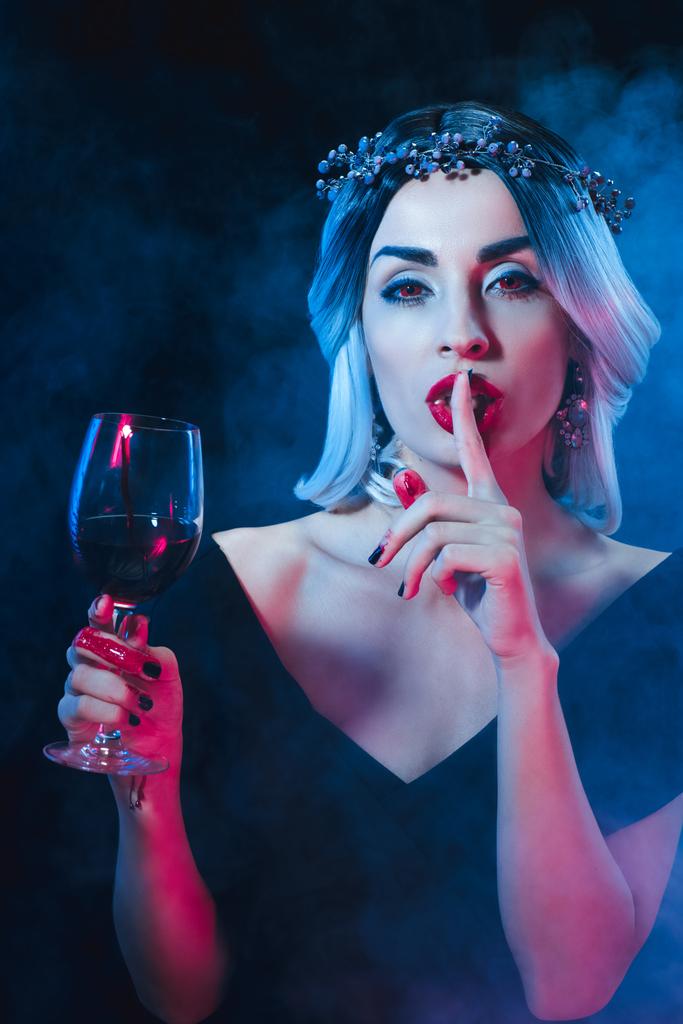 煙で暗い背景に沈黙の記号と血で持株のワイングラスを示すセクシーな吸血鬼の女  - 写真・画像